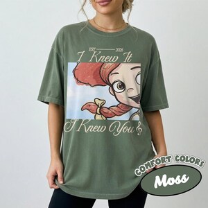 May include: A moss green short-sleeve t-shirt featuring a graphic of a cartoon character with red hair and a braid. The text reads EST 2026 I Knew It I Knew You in a serif font. The design is centered on the chest of the garment.