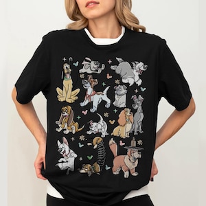 May include: Black t-shirt featuring a colorful cartoon illustration of various dog breeds. The design includes characters like Pluto, Lady, Tramp, and others, with floral and heart accents. The shirt has a white crew neck collar.