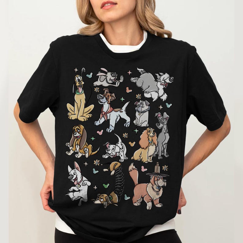 May include: Black t-shirt featuring a colorful cartoon illustration of various dog breeds. The design includes characters like Pluto, Lady, Tramp, and others, with floral and heart accents. The shirt has a white crew neck collar.