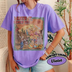 Op de afbeelding: Een violetkleurig T-shirt met korte mouwen en een grafische print met de tekst I KNEW IT, I KNEW YOU. De illustratie toont een groep geanimeerde personages in een woestijnomgeving met cactussen, een gitaar en een ster-vormige sheriff-badge.