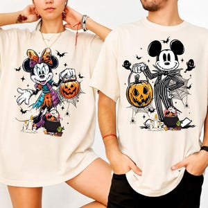 May include: Beige t-shirts featuring Halloween-themed cartoon characters. One shirt displays Minnie Mouse in a witch costume holding a jack-o'-lantern. The other shirt shows Mickey Mouse dressed as Jack Skellington, also with a pumpkin.