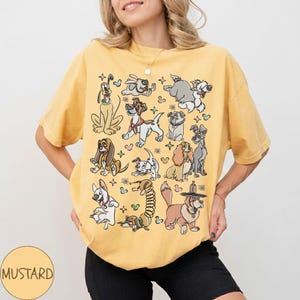 May include: Mustard-colored t-shirt featuring a collection of cartoon dog illustrations. The dogs are in various poses and styles, with a few floral and star accents. The shirt is a relaxed fit.