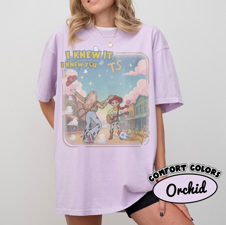 May include: An orchid-colored t-shirt featuring a graphic print of two cartoon characters playing guitars in a western town setting. The text "I KNEW IT, I KNEW YOU TS" is printed above the characters. The design includes clouds, stars, and a sheriff star.