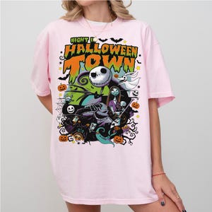 May include: Pink t-shirt featuring a colorful graphic from "The Nightmare Before Christmas." The design includes Jack Skellington, Sally, and other characters, with the words "Nightmare Halloween Town" in orange and green.