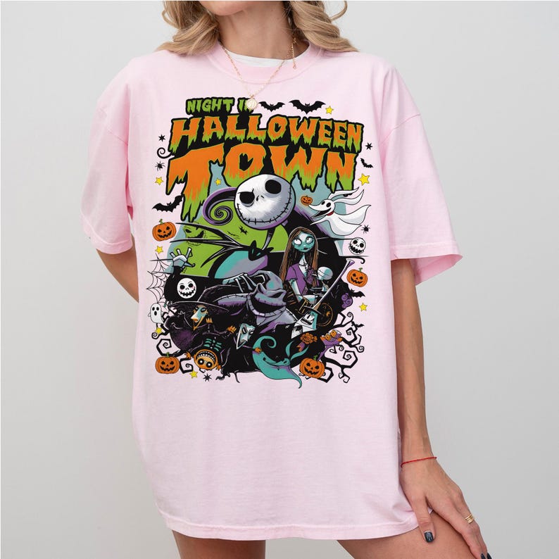 May include: Pink t-shirt featuring a colorful graphic from "The Nightmare Before Christmas." The design includes Jack Skellington, Sally, and other characters, with the words "Nightmare Halloween Town" in orange and green.