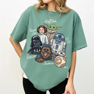 May include: Sage green t-shirt with a graphic of Star Wars characters: Darth Vader, Princess Leia, Baby Yoda, Chewbacca, R2-D2, and BB-8. The names of the characters are written in a handwritten font.