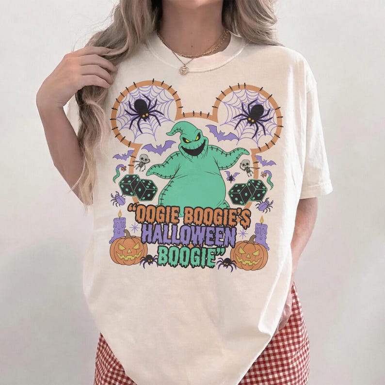 May include: Cream-colored t-shirt featuring a green Oogie Boogie character design. The design includes spiders, dice, bats, and pumpkins, with the text "Oogie Boogie's Halloween Boogie" in a purple and green font. The shirt has a round neckline and short sleeves.