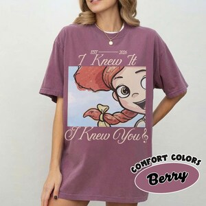 May include: A berry-colored short-sleeve t-shirt featuring a graphic print of a cartoon character with red hair and a musical note. The text reads EST 2026, I Knew It, and I Knew You. The shirt has a crew neckline and a relaxed, loose-fitting silhouette.
