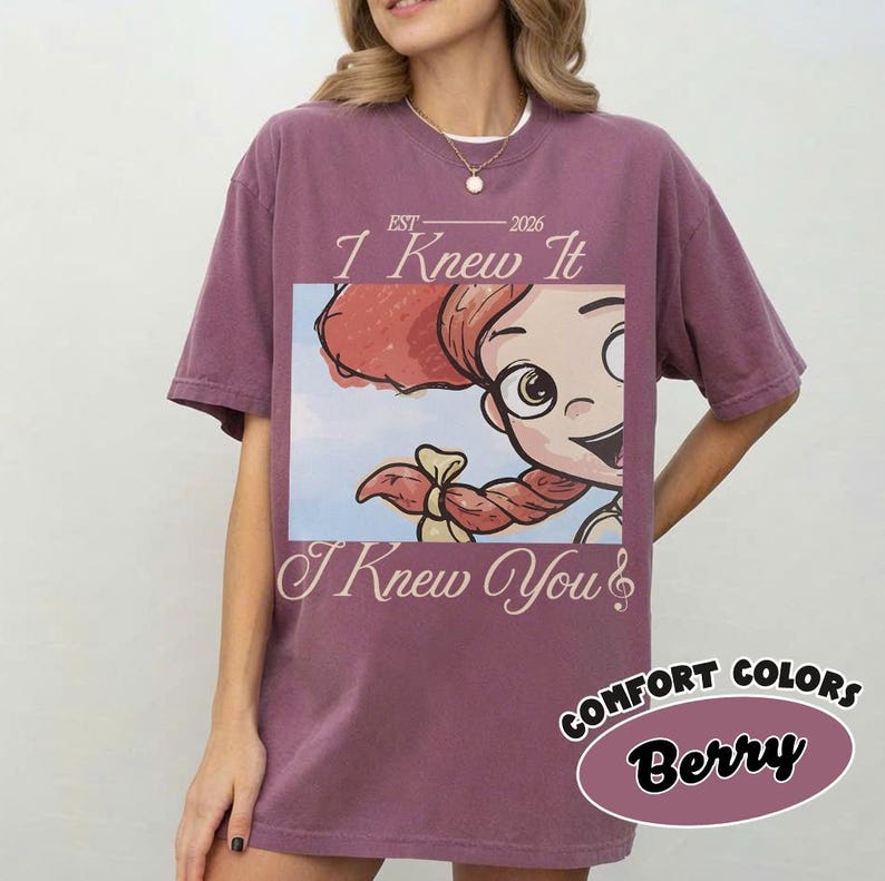 May include: A berry-colored short-sleeve t-shirt featuring a graphic print of a cartoon character with red hair and a musical note. The text reads EST 2026, I Knew It, and I Knew You. The shirt has a crew neckline and a relaxed, loose-fitting silhouette.