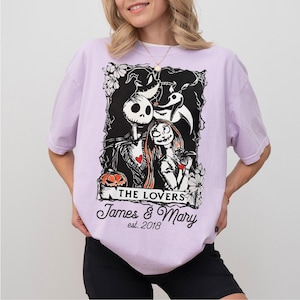 May include: A light purple t-shirt featuring a black and white graphic of Jack Skellington and Sally from The Nightmare Before Christmas. The graphic includes the text "THE LOVERS" and personalized names and date.
