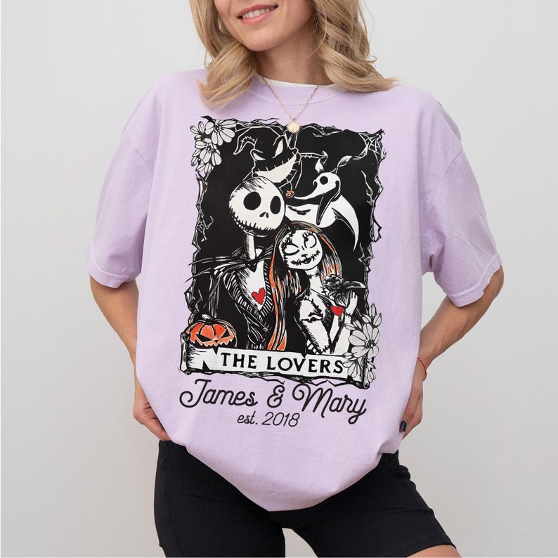 May include: A light purple t-shirt featuring a black and white graphic of Jack Skellington and Sally from The Nightmare Before Christmas. The graphic includes the text "THE LOVERS" and personalized names and date.
