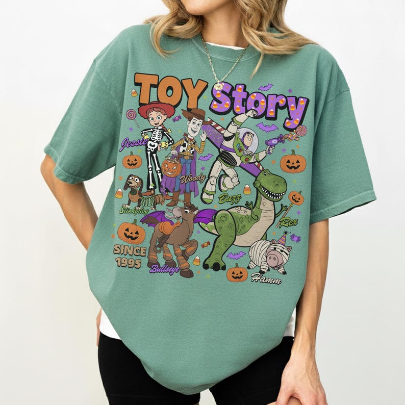 Op de afbeelding: Een saliegroen t-shirt met een Toy Story Halloween-afbeelding met de personages Jessie, Woody, Buzz Lightyear, Slinky Dog, Bullseye, Rex en Hamm. Het ontwerp bevat pompoenen, maissnoepjes, vleermuizen en de tekst "TOY STORY" en "SINCE 1995".