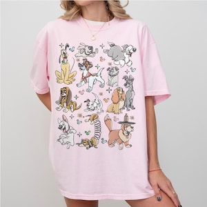 May include: A light pink t-shirt featuring a collection of cartoon dog illustrations. The dogs are in various poses and styles, with black outlines and colorful accents. The shirt has short sleeves and a relaxed fit.