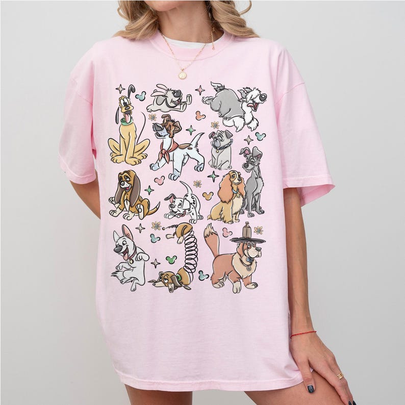 May include: A light pink t-shirt featuring a collection of cartoon dog illustrations. The dogs are in various poses and styles, with black outlines and colorful accents. The shirt has short sleeves and a relaxed fit.