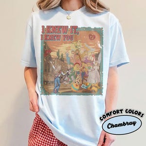Op de afbeelding: Een lichtblauw t-shirt met korte mouwen en een grafische print met de tekst I KNEW IT, I KNEW YOU. Het ontwerp toont een groep geanimeerde figuren in een woestijnomgeving met cactussen, een bal en een sheriffster binnen een decoratieve rand.