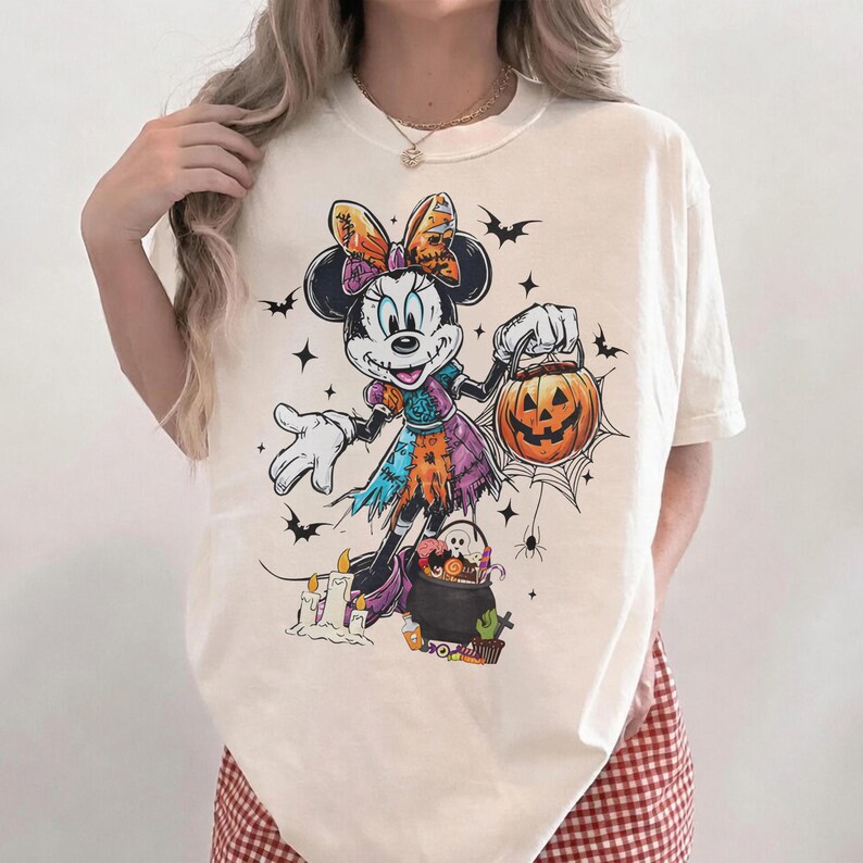 May include: Cream-colored t-shirt featuring a Halloween-themed Minnie Mouse graphic. Minnie is dressed in a patchwork dress and holding a jack-o'-lantern. Bats, stars, and spiderwebs surround the character, creating a festive design.