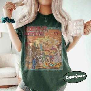 Op de afbeelding: Een donkergroen t-shirt met een grafische print met de tekst "I KNEW IT, I KNEW YOU" boven een woestijnsc&egrave;ne met meerdere ge&iuml;llustreerde figuren, waaronder een persoon die gitaar speelt, en een klein hartje met de letters "TS".