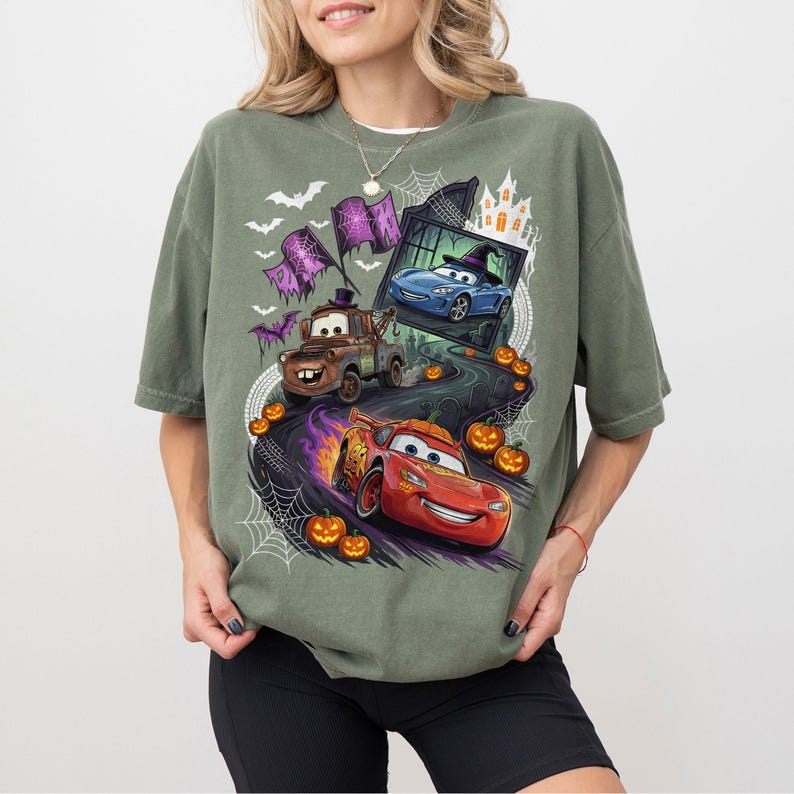 K&ouml;nnte beinhalten: Ein olivgr&uuml;nes T-Shirt mit einem Halloween-Motiv, das Zeichentrickautos zeigt, darunter einen braunen Abschleppwagen und einen roten Rennwagen. Das Design umfasst K&uuml;rbisse, Spinnennetze, Flederm&auml;use und die Silhouette eines Spukhauses auf einer dunklen Stra&szlig;e.