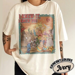 Op de afbeelding: Een ivoorkleurig t-shirt met korte mouwen en een grafische print met de tekst "I KNEW IT, I KNEW YOU" en "TS". De illustratie toont een groep geanimeerde figuren in een woestijnomgeving met een gitaar, een bal en een sheriffster.