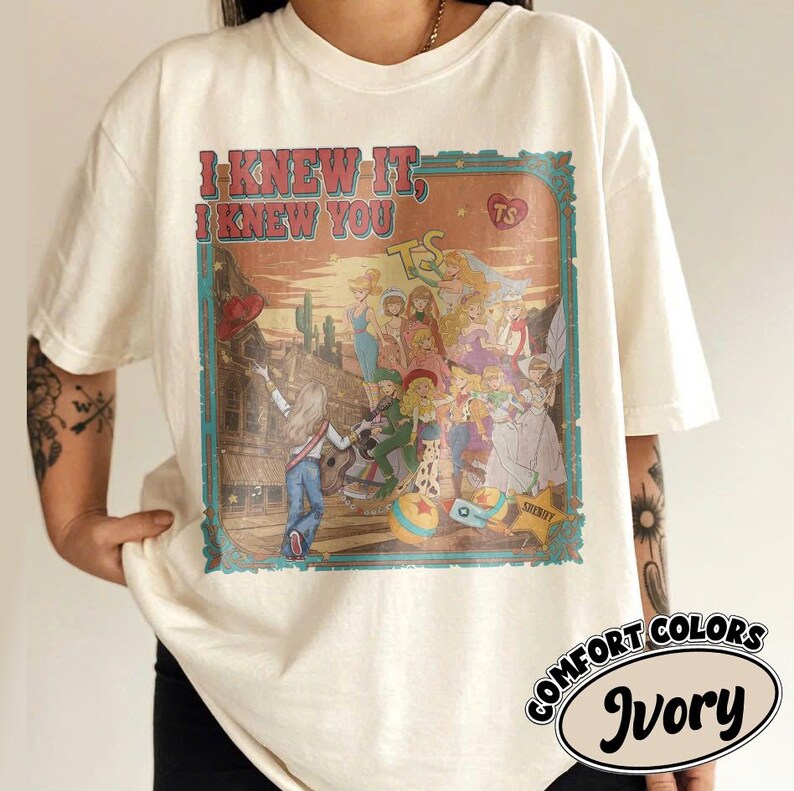 Op de afbeelding: Een ivoorkleurig t-shirt met korte mouwen en een grafische print met de tekst "I KNEW IT, I KNEW YOU" en "TS". De illustratie toont een groep geanimeerde figuren in een woestijnomgeving met een gitaar, een bal en een sheriffster.
