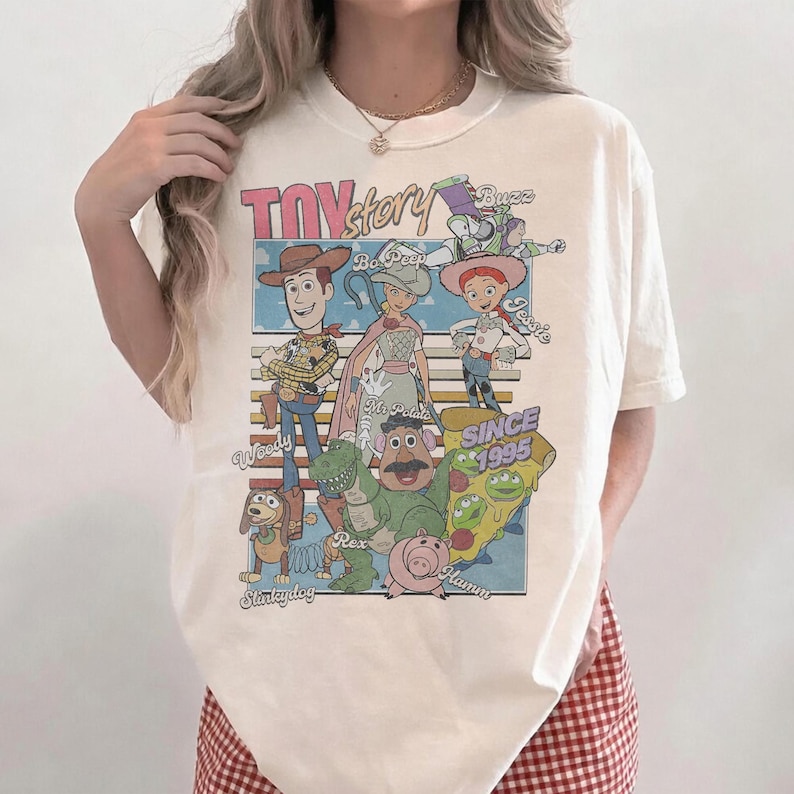 Pu&ograve; includere: T-shirt color crema con grafica dei personaggi di Toy Story, tra cui Woody, Buzz Lightyear e Jessie. Il design include il testo "Toy Story" e "Since 1995". La maglietta ha un girocollo classico e maniche corte.