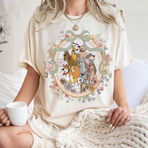 May include: Cream-colored t-shirt featuring a colorful illustration of various cartoon dogs within an oval frame. The frame is adorned with flowers and a green ribbon. The shirt has short sleeves and a relaxed fit.