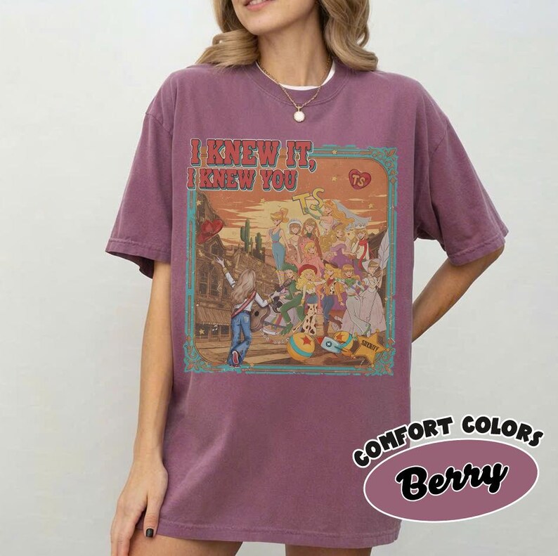 Op de afbeelding: Een bessenkleurig T-shirt met korte mouwen en een rechthoekige grafische print met de tekst I KNEW IT, I KNEW YOU. De illustratie toont een groep geanimeerde personages in een woestijnomgeving met een zonsondergang, cactussen en een ster.