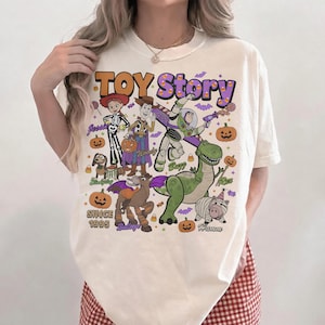 Op de afbeelding: Een cr&egrave;mekleurig t-shirt met een grafische print van Toy Story-personages zoals Jessie, Woody, Buzz Lightyear, Slinky Dog, Bullseye, Rex en Hamm in Halloween-kostuums. De tekst luidt Toy Story en Since 1995, omringd door pompoenen.