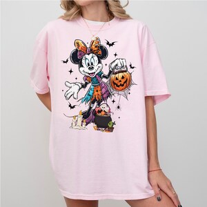May include: A light pink t-shirt featuring a Halloween-themed Minnie Mouse graphic. The cartoon Minnie Mouse is dressed in a witch costume and holding a jack-o'-lantern. Bats, stars, and a spiderweb complete the design.