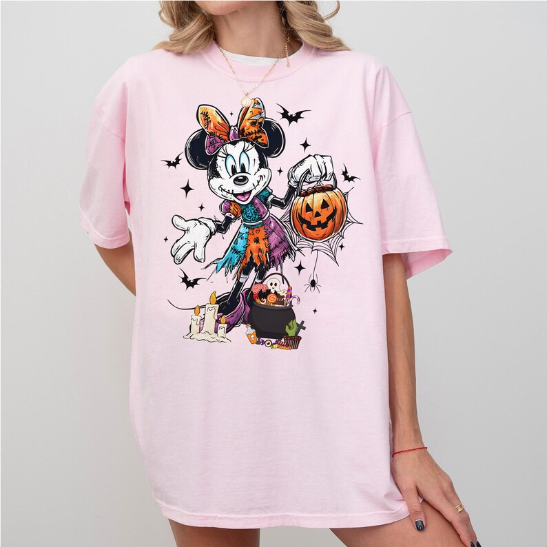 May include: A light pink t-shirt featuring a Halloween-themed Minnie Mouse graphic. The cartoon Minnie Mouse is dressed in a witch costume and holding a jack-o'-lantern. Bats, stars, and a spiderweb complete the design.