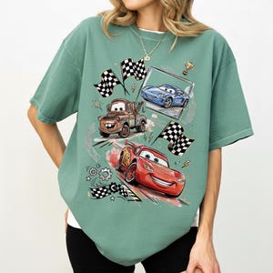 May include: A seafoam green t-shirt featuring a graphic of animated race cars, including a red car, a blue car, and a brown tow truck. The design includes checkered flags, a trophy, and speed lines, creating a racing theme.