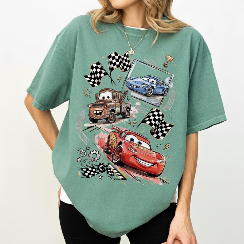 May include: A seafoam green t-shirt featuring a graphic of animated race cars, including a red car, a blue car, and a brown tow truck. The design includes checkered flags, a trophy, and speed lines, creating a racing theme.