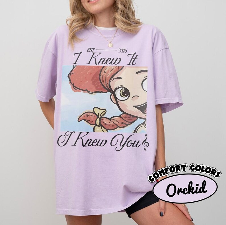 May include: A light purple short-sleeve t-shirt featuring a graphic of a cartoon character with red hair and a braid. The text reads EST 2026, I Knew It, and I Knew You with a musical note symbol. The shirt has a crew neckline and a relaxed fit.
