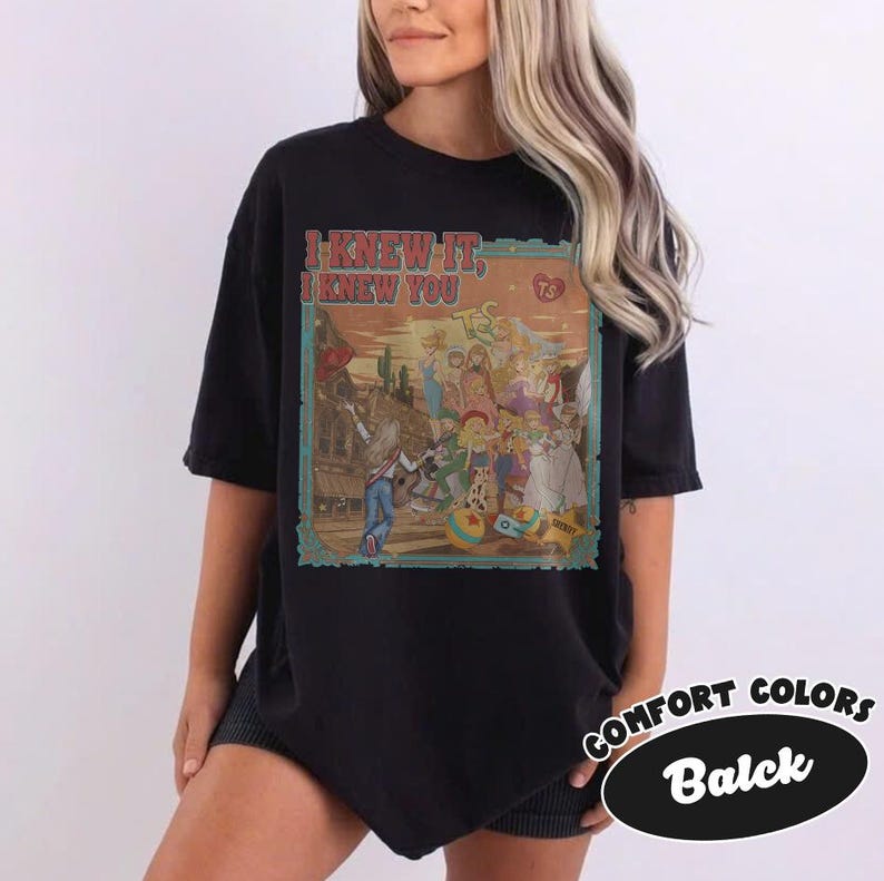 Op de afbeelding: Een zwart t-shirt met korte mouwen en een rechthoekige grafische print met de tekst I KNEW IT, I KNEW YOU. De illustratie toont een groep geanimeerde personages in een woestijnomgeving met een cactus, een bal en een stervormige badge.