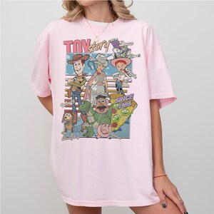 Pu&ograve; includere: T-shirt rosa chiaro con una grafica dei personaggi di Toy Story, tra cui Woody, Buzz Lightyear e Jessie. Il design include il testo "Toy Story" e "Since 1995". La maglietta ha maniche corte e girocollo.