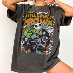 May include: Dark gray t-shirt featuring a colorful graphic from "The Nightmare Before Christmas." The design includes Jack Skellington, Sally, and other characters, with the text "Night in Halloween Town" in orange and green.