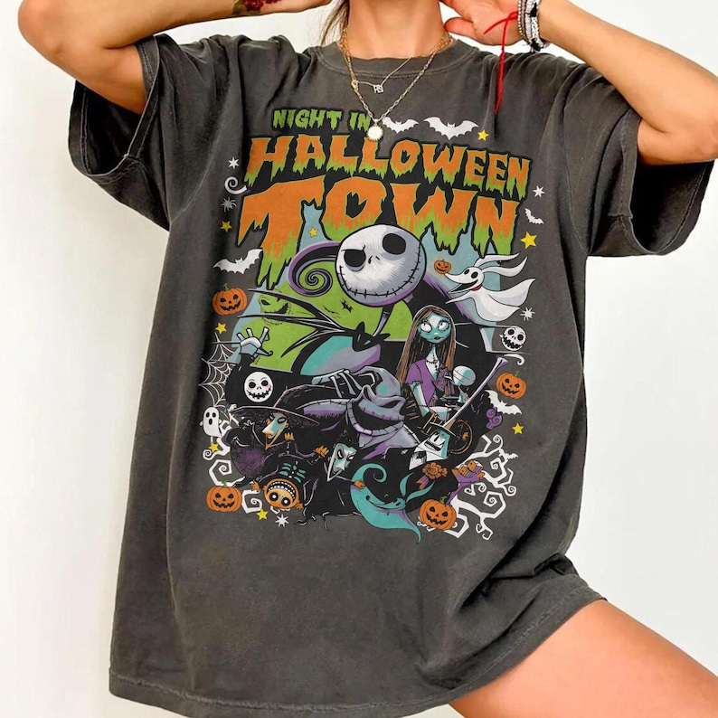 May include: Dark gray t-shirt featuring a colorful graphic from "The Nightmare Before Christmas." The design includes Jack Skellington, Sally, and other characters, with the text "Night in Halloween Town" in orange and green.