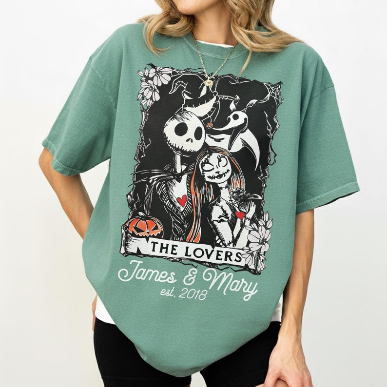 May include: A green t-shirt featuring a graphic of Jack Skellington and Sally from The Nightmare Before Christmas, with the words "THE LOVERS" and "James & Mary est. 2018". The design includes a pumpkin and floral accents.