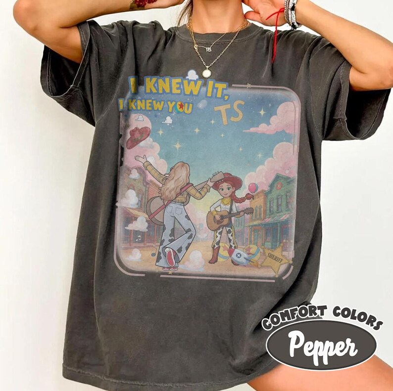 May include: A charcoal gray short-sleeve t-shirt featuring a graphic print of two characters in a western town setting. The text "I KNEW IT, I KNEW YOU TS" is printed above the characters. The design includes a starry sky, clouds, and guitars.