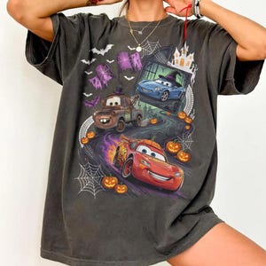 K&ouml;nnte beinhalten: Ein anthrazitfarbenes T-Shirt mit einem Grafikdruck von Charakteren aus dem Film Cars in einer Halloween-Umgebung. Das Design zeigt ein rotes Rennauto, einen braunen Abschleppwagen, ein blaues Auto, K&uuml;rbisse, Spinnennetze, Flederm&auml;use und ein Spukhaus.