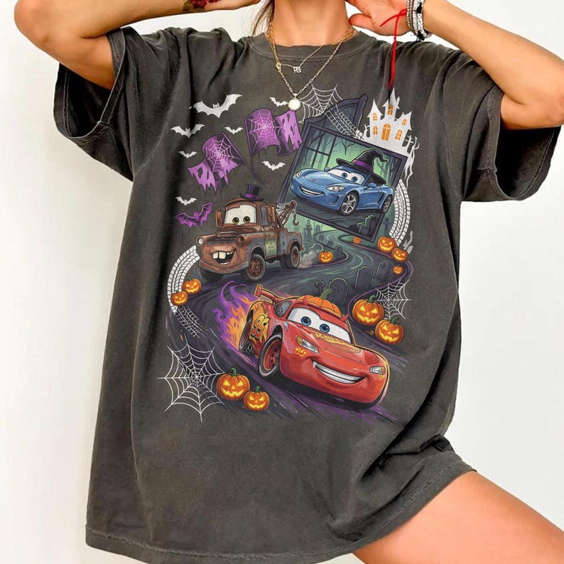 K&ouml;nnte beinhalten: Ein anthrazitfarbenes T-Shirt mit einem Grafikdruck von Charakteren aus dem Film Cars in einer Halloween-Umgebung. Das Design zeigt ein rotes Rennauto, einen braunen Abschleppwagen, ein blaues Auto, K&uuml;rbisse, Spinnennetze, Flederm&auml;use und ein Spukhaus.