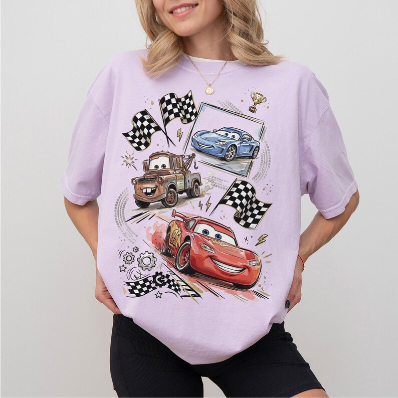 May include: A light purple t-shirt featuring a graphic of animated race cars, including a red car, a blue car, and a brown tow truck. Checkered flags and a trophy are also part of the design. The shirt is a casual style.