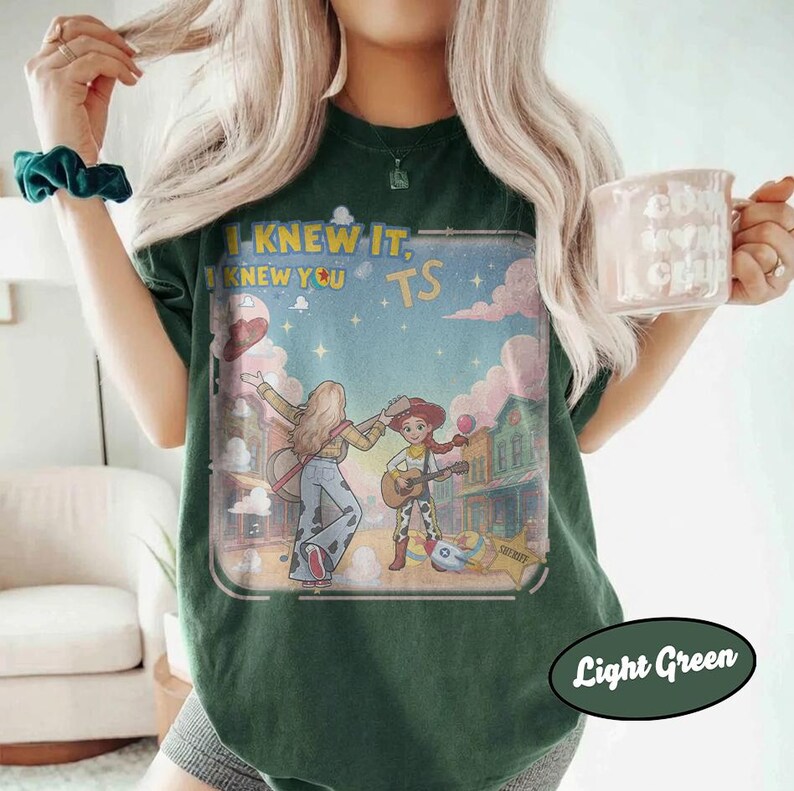 May include: A dark green t-shirt featuring a graphic print of two cartoon characters playing guitars in a western town setting. The text "I KNEW IT, I KNEW YOU" is printed above the characters. The shirt has a crew neckline and short sleeves.