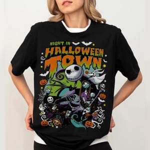 May include: Black t-shirt featuring a colorful graphic from "The Nightmare Before Christmas." The design includes Jack Skellington, Sally, Zero, and other characters, with the text "Night in Halloween Town."