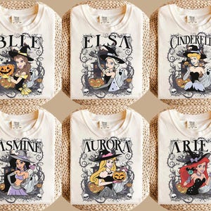 May include: Six off-white t-shirts featuring graphic prints of cartoon characters dressed as witches for Halloween. Each shirt displays a character name, including Belle, Elsa, Cinderella, Jasmine, Aurora, and Ariel, surrounded by spooky vines, ghosts, and pumpkins.