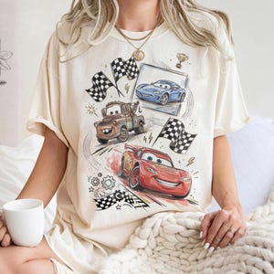 May include: Cream-colored t-shirt featuring a colorful graphic of animated race cars, including a red car, a blue car, and a brown tow truck. The design includes checkered flags, lightning bolts, and gears.