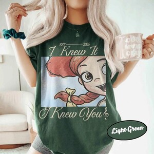 May include: A dark green short-sleeve t-shirt featuring a graphic of a cartoon character with red hair in a ponytail. The text reads EST 2026 I Knew It I Knew You. The shirt has a crew neckline and a relaxed fit, showcasing a vintage-inspired design.