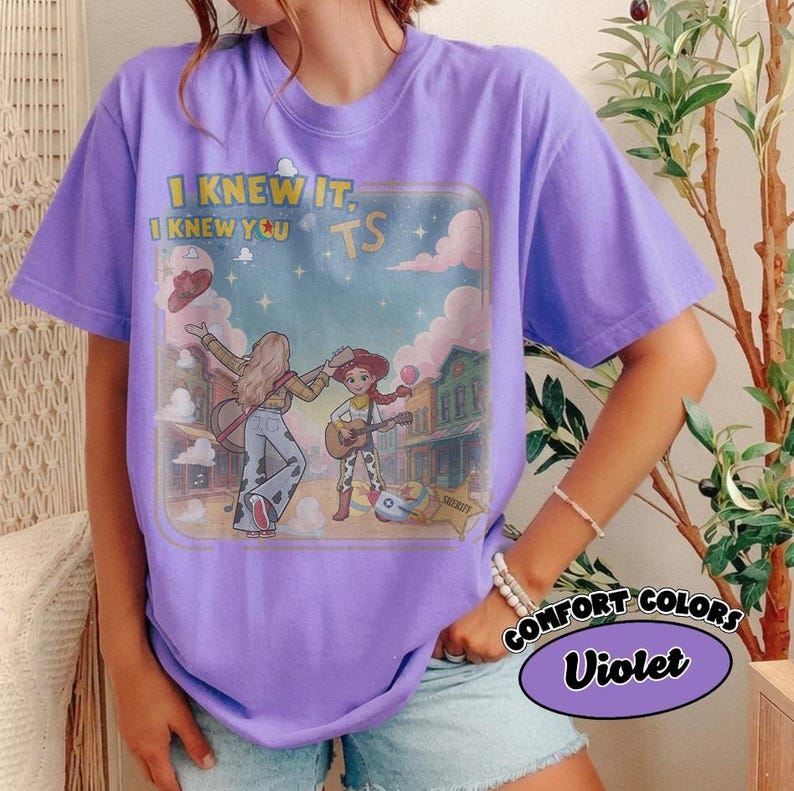 May include: A violet t-shirt featuring a graphic print of a person and a cowgirl character playing guitars in a western town setting. The text reads I KNEW IT, I KNEW YOU TS. The design includes clouds, stars, a sheriff badge, and a hat.