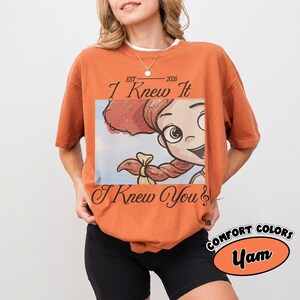 May include: A burnt orange short-sleeve t-shirt featuring a graphic of a cartoon character with red hair in braids. The text reads "EST 2026 I Knew It I Knew You" in a black script font. The shirt has a crew neckline and a relaxed fit.