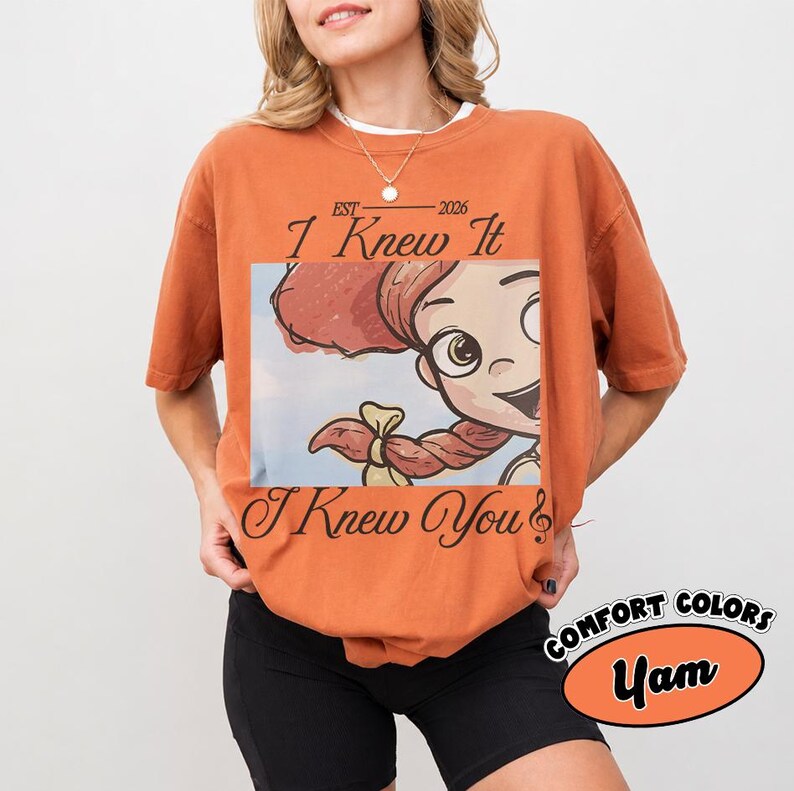 May include: A burnt orange short-sleeve t-shirt featuring a graphic of a cartoon character with red hair in braids. The text reads "EST 2026 I Knew It I Knew You" in a black script font. The shirt has a crew neckline and a relaxed fit.